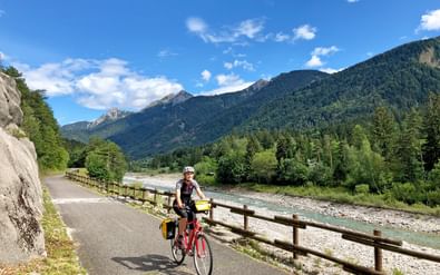 Fietser met rode fiets op geasfalteerd pad langs rivier met houten hek, omringd door groene bossen en bergen onder blauwe lucht.