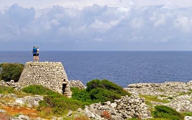 Twee wandelaars staan op oude stenen ruïnes met uitzicht op de blauwe zee bij Marina Serra. Mediterrane vegetatie en rotsachtige kust zichtbaar.