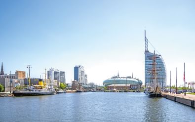 Haventafereel met historische zeilschepen aangemeerd voor moderne gebouwen. Heldere blauwe lucht weerkaatst op kalm water.