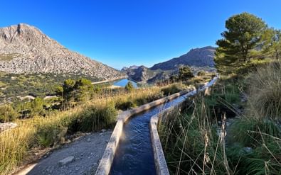 Stenen waterkanaal naar Gorg Blau reservoir in Mallorca's Tramuntana gebergte, omgeven door mediterrane vegetatie en rotsachtige toppen.