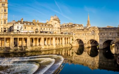 Pulteney Bridge met drie stenen bogen over de rivier de Avon in Bath. Historische honingkleurige gebouwen langs de oever onder een blauwe lucht.