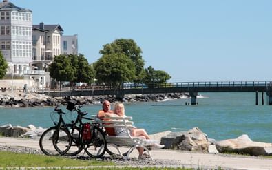 Twee fietsers zitten op een bankje aan de Oostzee met hun fietsen dichtbij. Witte gebouwen en een pier op de achtergrond.