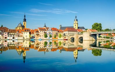 Historische stenen brug over de Main in Kitzingen met kleurrijke gebouwen en kerktorens weerspiegeld in kalm water onder blauwe lucht.