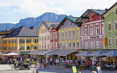 Stadskern Mondsee met kleurrijke historische gebouwen in geel, rood en groen. Mensen dineren op buitenterrassen op het plein met bergen op de achtergrond.