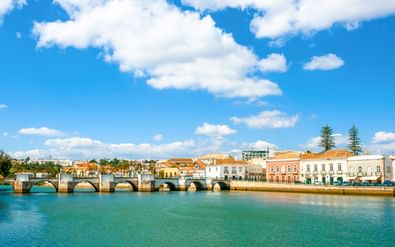 Stenen brug met meerdere bogen over een rivier in Tavira, Algarve. Kleurrijke historische gebouwen langs de waterkant onder een blauwe lucht.