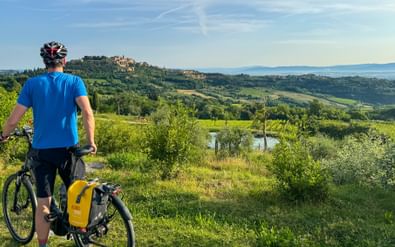 Fietser in blauw shirt met helm staat naast fiets met gele tassen, kijkend naar heuveltop stad Montepulciano over groen Toscaans landschap.