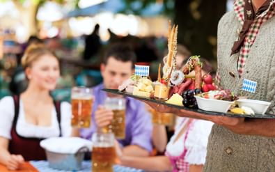 Ober in traditioneel vest houdt houten plank met kaas, pretzels en snacks. Gasten in Beierse klederdracht met bierpullen zitten aan tafel op de achtergrond.