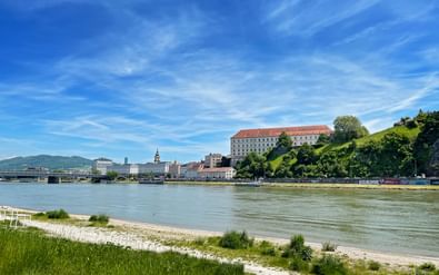Uitzicht over de Donau naar het Kasteel Museum Linz, een groot barok gebouw op een heuvel, met de stadshorizon en bergen op de achtergrond.
