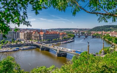 Panoramisch uitzicht op Praag met historisch burchtcomplex, rode pannendaken en stenen bruggen over de Moldau, omlijst door groen gebladerte.