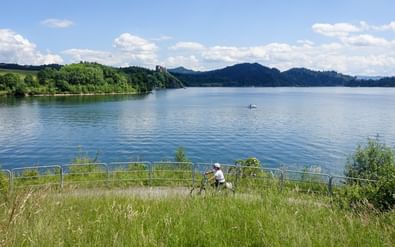 Fietser op grasrijke oever van Czorsztyn-stuwmeer met blauw water, beboste heuvels en bergen onder gedeeltelijk bewolkte hemel.