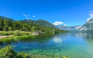 Kristalhelder turquoise meer omgeven door beboste bergen in de Julische Alpen. Een houten steiger steekt het water in met gebouwen zichtbaar aan de oever.
