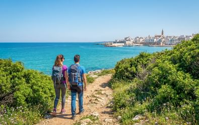 Twee wandelaars met rugzakken op een kustpad in Apulië, met uitzicht op turquoise zee en historische stad met kerktoren.