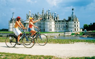 Twee fietsers met fietsen poseren voor een sierlijk Frans Renaissance-kasteel in de Loire-vallei met meerdere torens en schoorstenen.