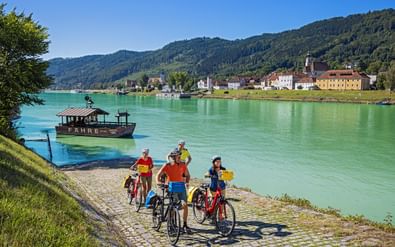 Vier fietsers met fietsen aan rivieroever wachten op veerboot in Engelhartszell. Groene Donau, beboste heuvels en kleurrijke gebouwen zichtbaar.