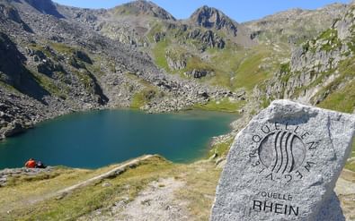 Stenen markering met 'Quelle Rhein' naast turquoise bergmeer omringd door rotsachtige bergen. Een rode tent is zichtbaar aan de oever.