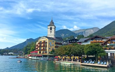 Meerzicht op St. Wolfgang met witte kerktoren, kleurrijke gebouwen en gele parasols op waterfront terras tegen bergachtergrond.
