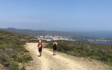 Twee wandelaars op een zandpad langs de Costa Brava kust met Middellandse Zee en witte kustplaats op de achtergrond.