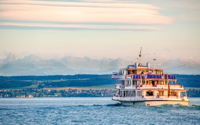 Witte passagiersveerboot met blauwe accenten vaart op de Bodensee bij Unteruhldingen. Bergen en kustlijn zichtbaar op de achtergrond onder avondlucht.