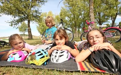 Vier kinderen met kleurrijke fietshelmen rusten op een deken tijdens een familiefietspauze, fietsen zichtbaar op de achtergrond tussen bomen.