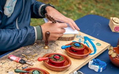 Handen die een pelgrimspas stempelen op de Portugese Camino. Houten stempels met waszegels liggen in terracotta schaaltjes op kleurrijke tafel.