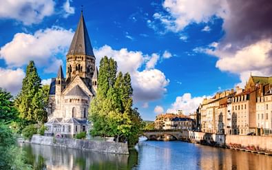Temple Neuf kerk met hoge toren aan de Moezel in Metz, met historische gebouwen en stenen brug onder blauwe lucht met witte wolken.