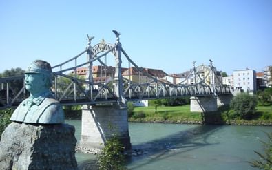 Historische stalen vakwerkbrug over de Salzach in Oberndorf met bronzen standbeeld op voorgrond en kleurrijke gebouwen op achtergrond.
