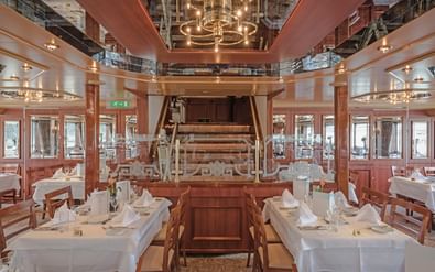 Elegant restaurant aan boord van MS Switzerland met wit gedekte tafels, houten stoelen en centrale piano. Warme verlichting en ramen aan beide zijden.