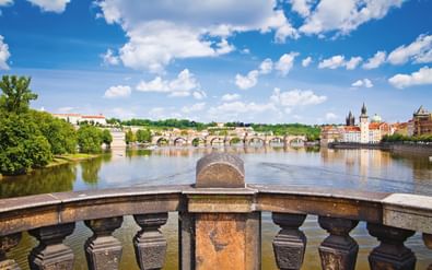 Uitzicht op Praags Karelsbrug over de Moldau, met historische gebouwen en torens zichtbaar op de rechteroever onder blauwe lucht.