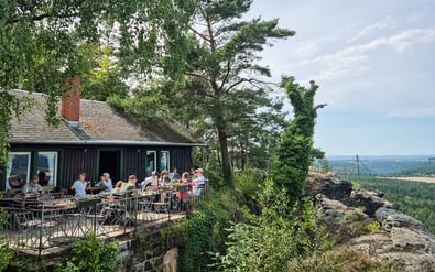 Buitenterras van een houten restaurant op een rotsachtige klif met gasten die dineren. Omringd door bomen met panoramisch uitzicht over boslandschap.