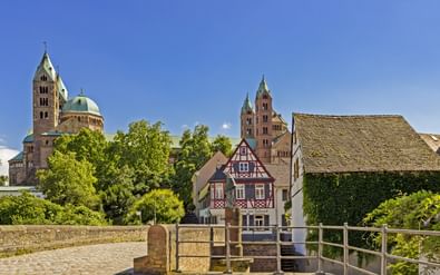 Zicht op de Dom van Speyer met dubbele torens en groene koepel achter traditionele vakwerkhuizen onder blauwe hemel.