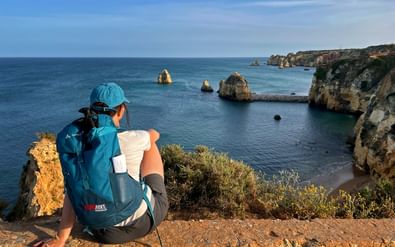 Wandelaar met blauwe rugzak zittend op kliprand met uitzicht op dramatische gouden kalksteenkliffen en turquoise water bij Ponta da Piedade.