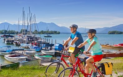 Twee fietsers met helmen op rode fietsen bij het Chiemsee meer. Jachthaven met boten, Herreninsel eiland en bergen op de achtergrond.
