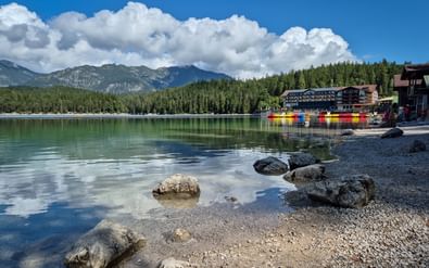 Rotsachtige oever van het Eibsee-meer met helder turquoise water, kleurrijke boten en gebouwen. Dicht bos en Zugspitze onder blauwe lucht.