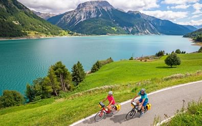 Twee fietsers op een geasfalteerde weg naast een turquoise alpenmeer omgeven door groene bergen en bossen onder een deels bewolkte hemel.