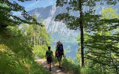 Twee wandelaars met rugzakken op bospad met uitzicht op blauw meer omringd door bergen in Salzkammergut.