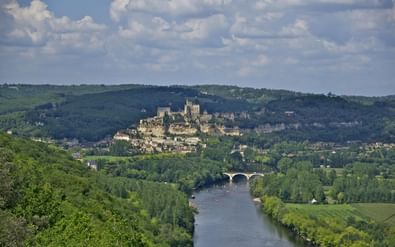 Kasteel Beynac op een heuvel met uitzicht op de Dordogne. Een stenen brug overspant de rivier, omgeven door groene bossen en heuvels.
