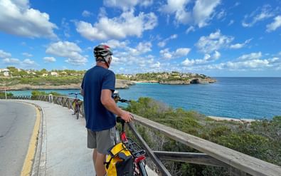 Fietser in blauw shirt en helm staat met fiets op kustpad met uitzicht op turquoise Middellandse Zee en Porto Cristo kustlijn.