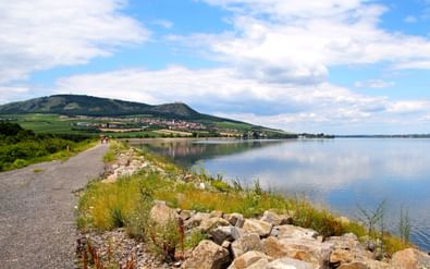 Grindfietspad langs een kalm meer in Pálava, Tsjechië, met rotsachtige oever, groene heuvels en dorp in de verte onder blauwe lucht.