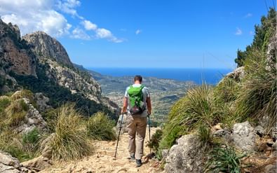 Wandelaar met groene rugzak op rotsachtig bergpad op Mallorca, uitkijkend over Middellandse Zee en kalkstenen kliffen.
