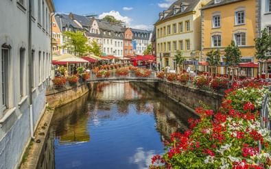 Historisch stadscentrum van Saarburg met stenen brug over kanaal met rode en witte bloemen. Kleurrijke gebouwen en terrasjes omringen de waterweg.