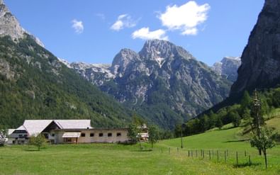 Bergdal in Lepena, Slovenië met traditionele alpenhuizen op groene weiden omringd door dramatische kalksteenpieken onder blauwe lucht.