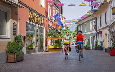 Twee fietsers rijden door een voetgangersstraat in Villach versierd met kleurrijke paraplu's tussen historische gebouwen.