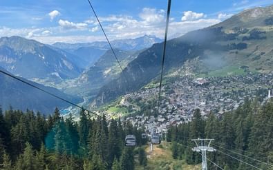 Kabelbaangondels dalen af naar Verbier gelegen in een Alpenvallei, omgeven door beboste bergen en toppen onder blauwe hemel.