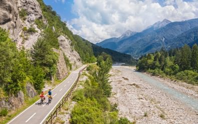 Twee fietsers op het Canal Valley fietspad dat door de bergen slingert. Het geasfalteerde pad loopt langs een rotsachtige rivierbedding met Alpentoppen.