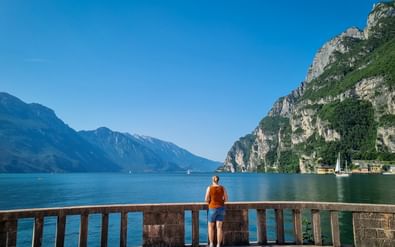Persoon in oranje trui staat bij stenen balustrade met uitzicht op Gardameer, omringd door dramatische bergen en helder blauw water.