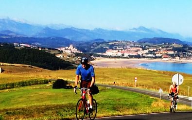 Twee fietsers op een kustweg met groene velden, een baai en dorpen op heuvels. Bergen zichtbaar op de achtergrond onder blauwe lucht.