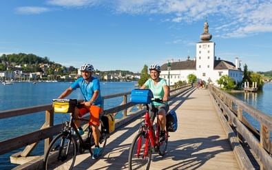 Twee fietsers met helmen en fietstassen pauzeren op houten brug naar kasteel Orth met witte toren aan meer in Salzkammergut.