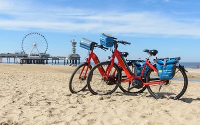 Twee huurfietsen geparkeerd op het zandstrand van Scheveningen met pier en reuzenrad zichtbaar op de achtergrond onder blauwe hemel met wolken.