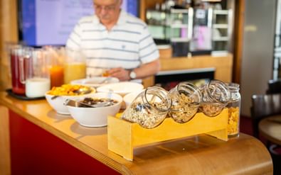 Ontbijtbuffet met graanpotjes in gele houder, kommen met fruit en yoghurt, sapkannen. Gast in gestreept shirt op achtergrond.