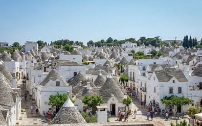 Panoramisch uitzicht op Alberobello met witte trulli-huizen met kenmerkende conische stenen daken. Toeristen lopen door met bomen omzoomde straten.
